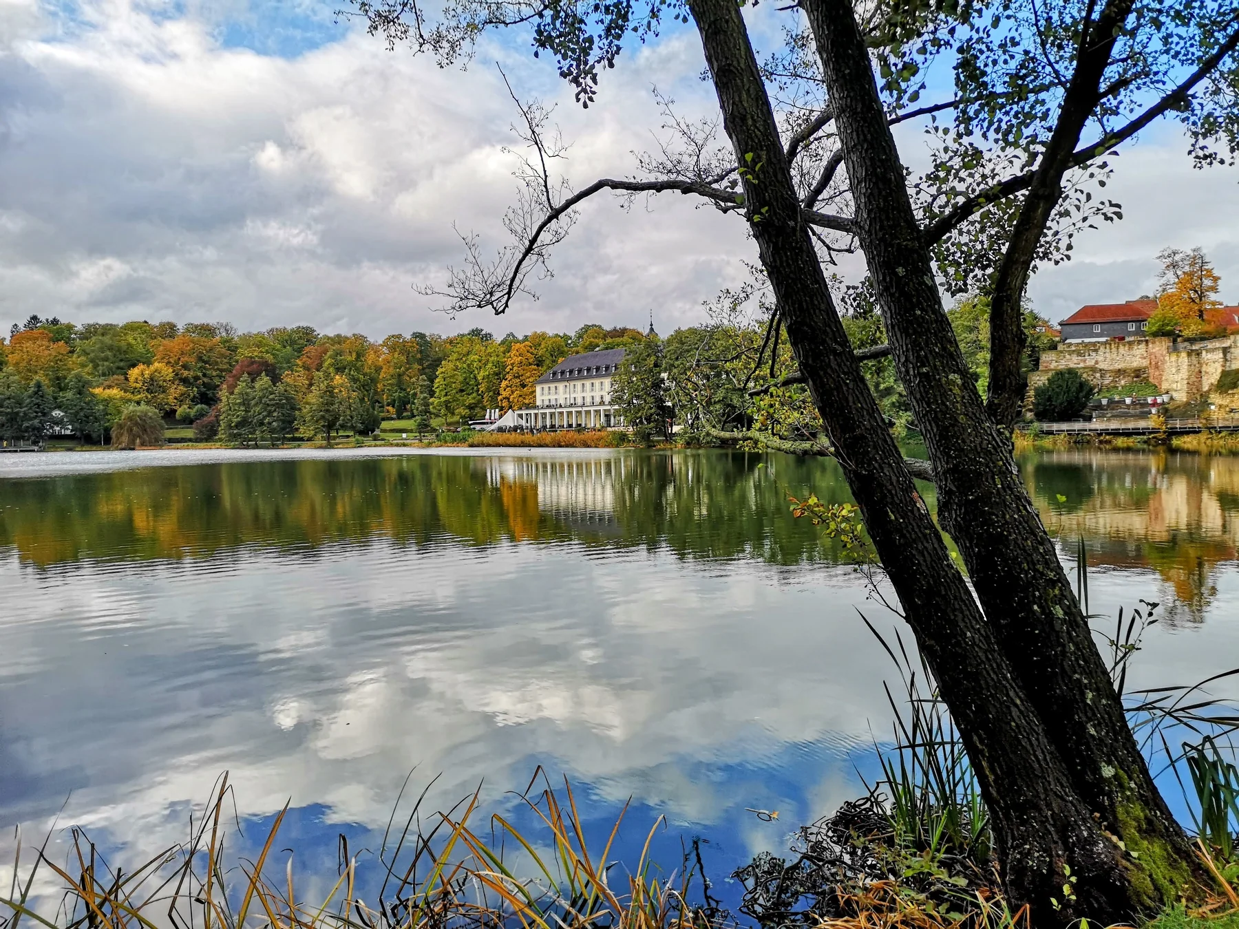 Herbstlicher Blick über den Burgsee im Rathenaupark in Bad Salzungen, mit Wolkenspiegelung im Wasser, buntem Laub und einem klassizistischen Gebäude am gegenüberliegenden Ufer