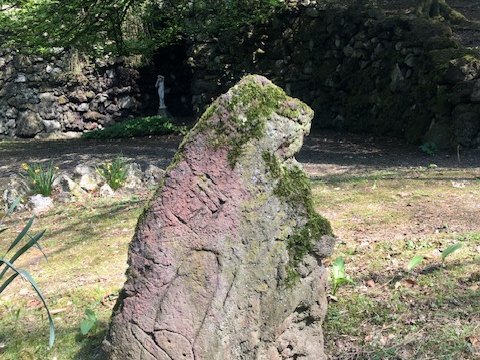 Großer Gesteinsblock auf einer Rasenfläche vor der bemoosten Bruchsteinmauer der Grotte