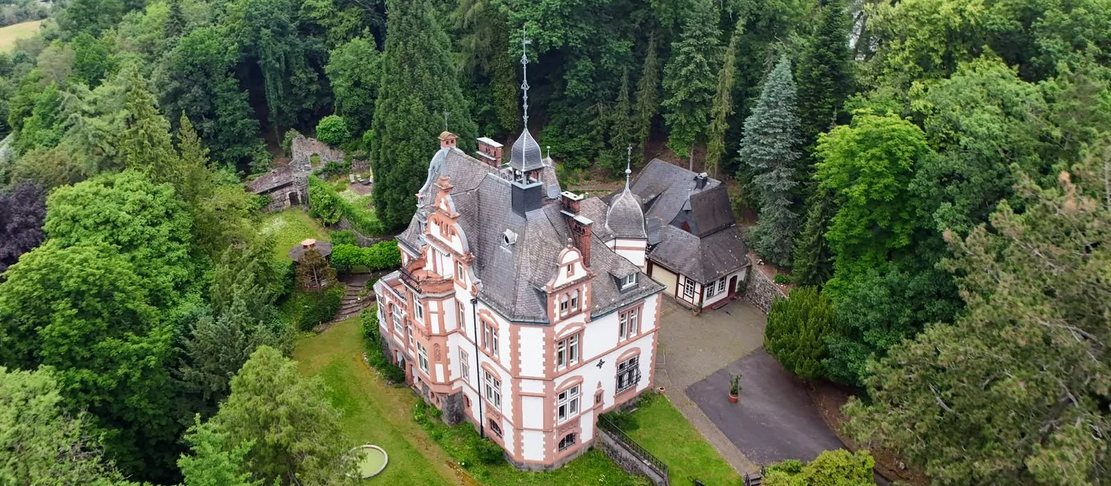 Luftaufnahme der Villa Haas in Sinn, das historistische Herrenhaus mit Turm und Fachwerk-Nebengebäude, ringsum der bewaldete Villenpark